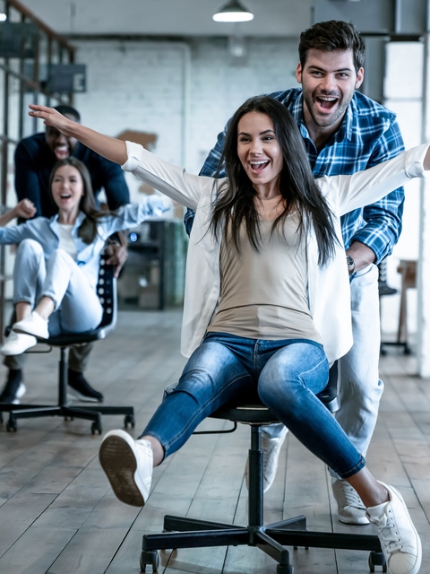 Work hard play hard. Four young cheerful business people in smart casual wear having fun while racing on office chairs and smiling.