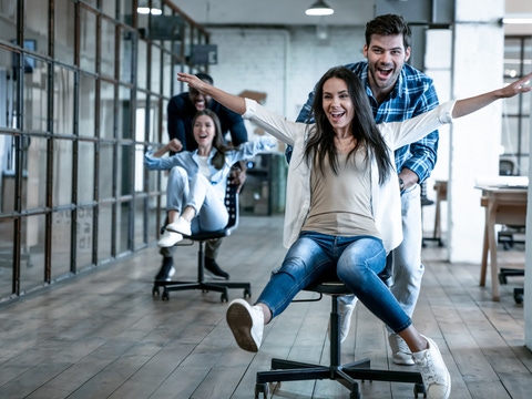 Work hard play hard. Four young cheerful business people in smart casual wear having fun while racing on office chairs and smiling.